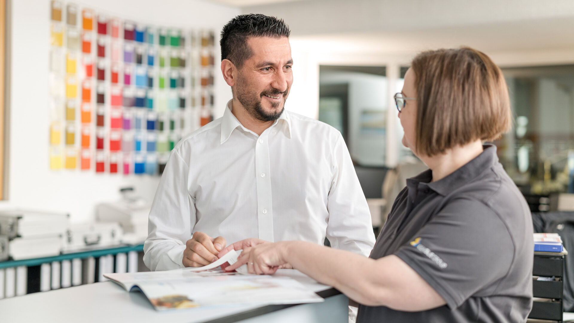 Beratungsgespräch zwischen zwei Mitarbeitenden im Büro, im Hintergrund eine Farbwand.