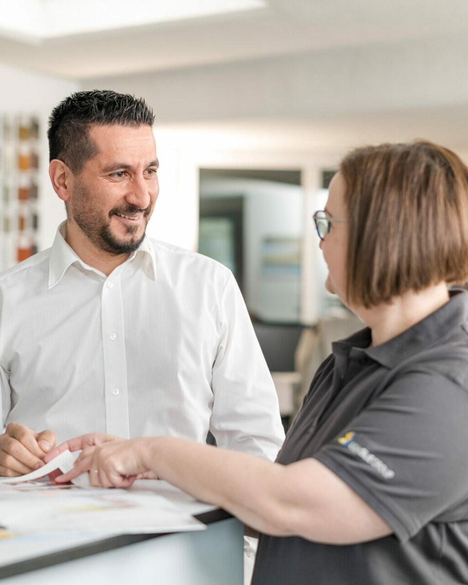 Beratungsgespräch zwischen zwei Mitarbeitenden im Büro, im Hintergrund eine Farbwand.