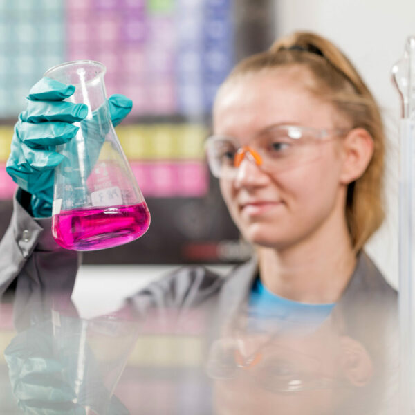 Laborantin mit Schutzbrille und Handschuhen hält ein Becherglas mit pinker Flüssigkeit vor einem Periodensystem.
