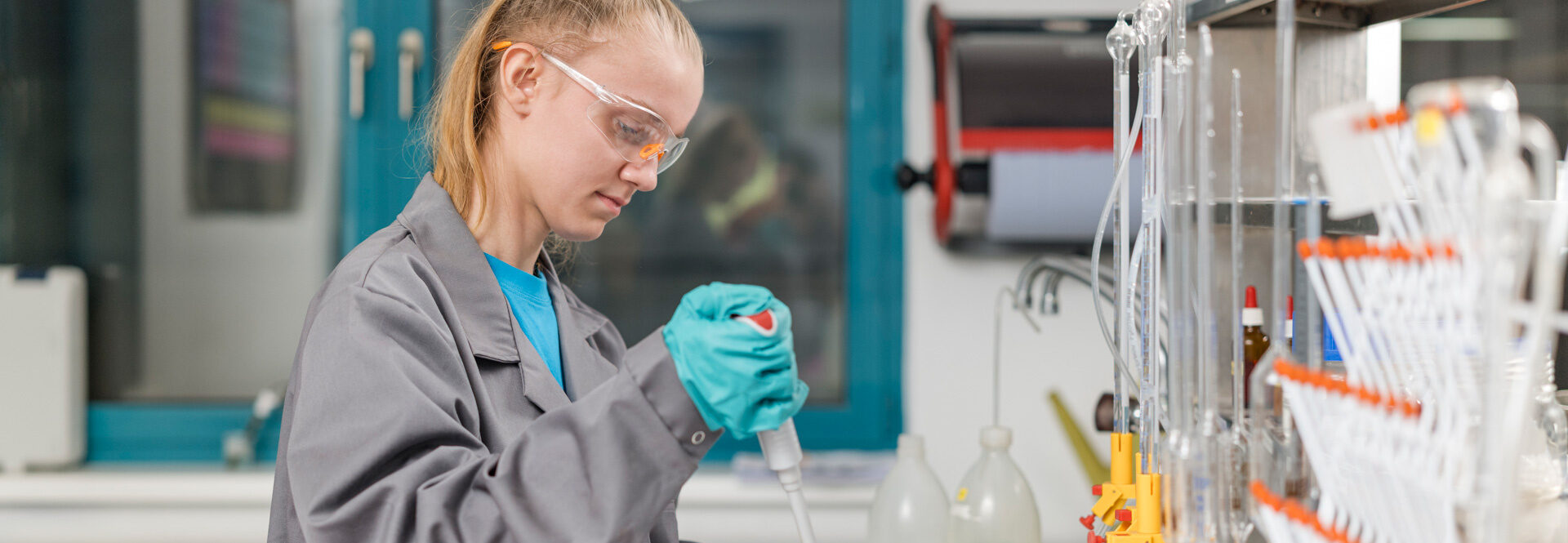 Laborantin in Schutzbrille und Handschuhen führt eine chemische Analyse mit einer Pipette in einem Becherglas durch.