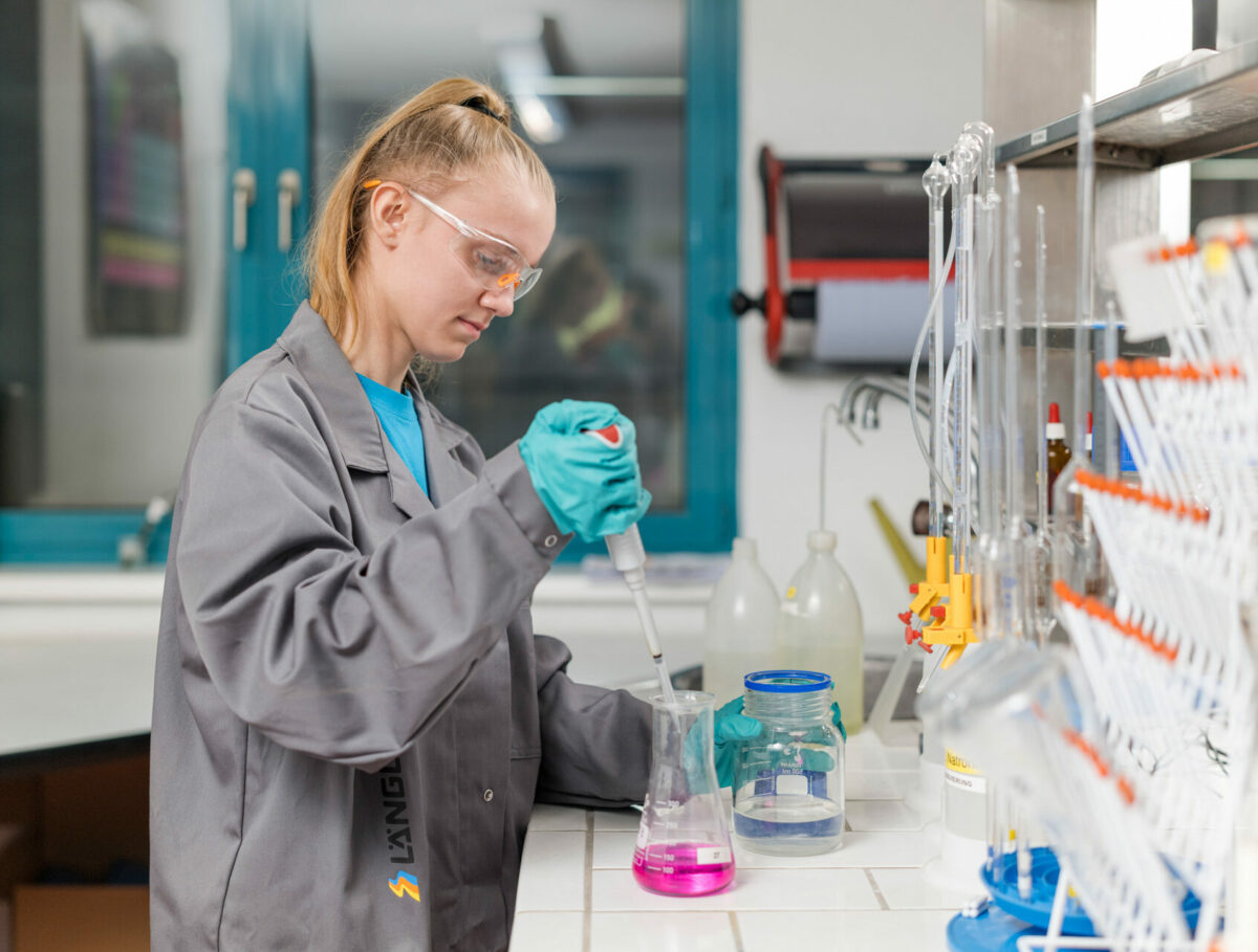 Laborantin in Schutzbrille und Handschuhen führt eine chemische Analyse mit einer Pipette in einem Becherglas durch.