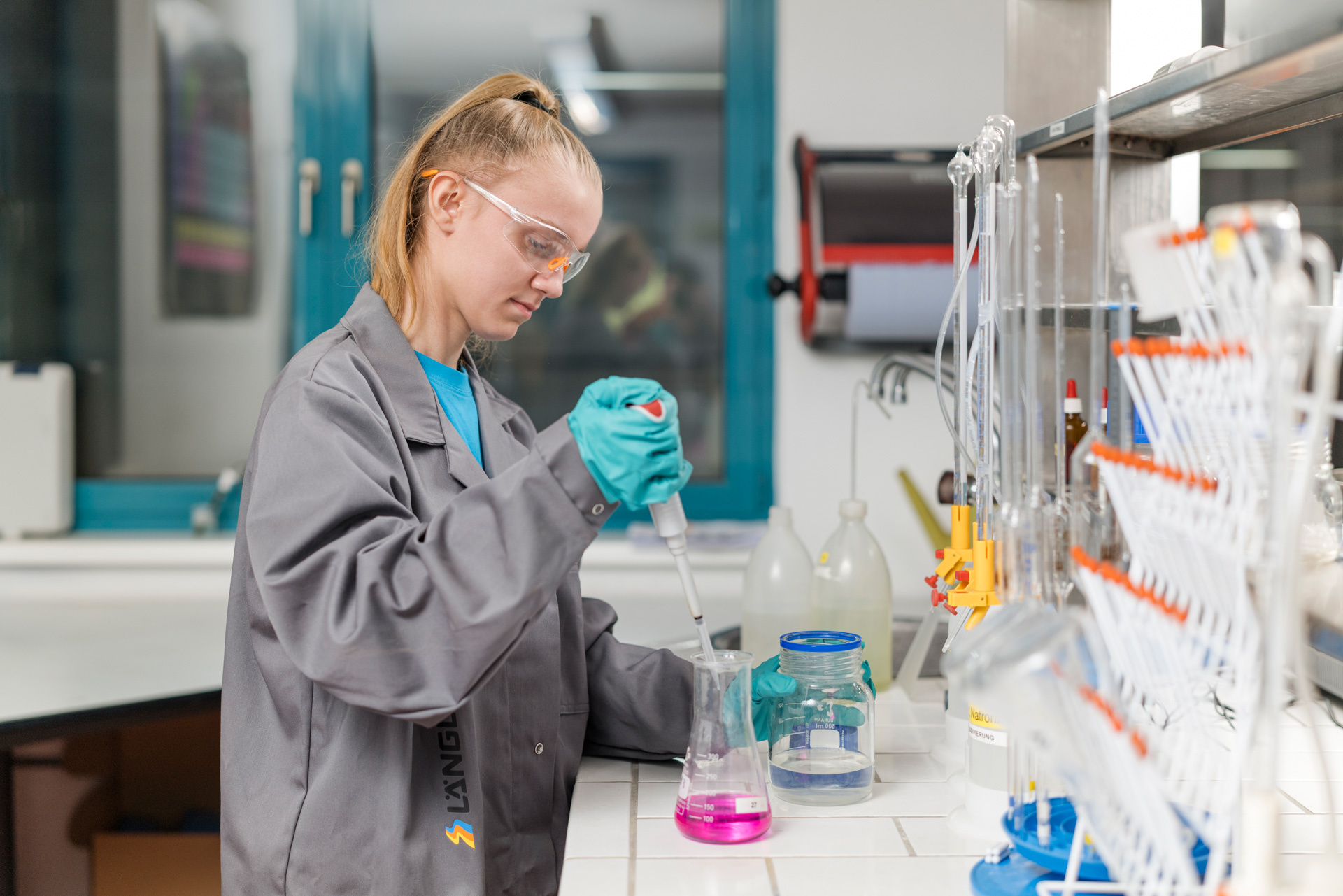 Laborantin in Schutzbrille und Handschuhen führt eine chemische Analyse mit einer Pipette in einem Becherglas durch.