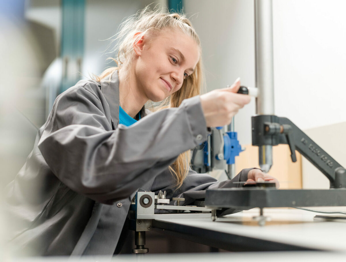 Eine junge Frau in grauer Arbeitskleidung führt eine Qualitätsprüfung an einer beschichteten Oberfläche durch.