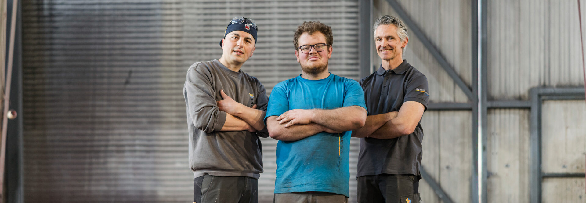 Drei Männer mit verschränkten Armen stehen in einer Industriehalle und blicken selbstbewusst in die Kamera. Sie tragen Arbeitskleidung und Sicherheitsschuhe.