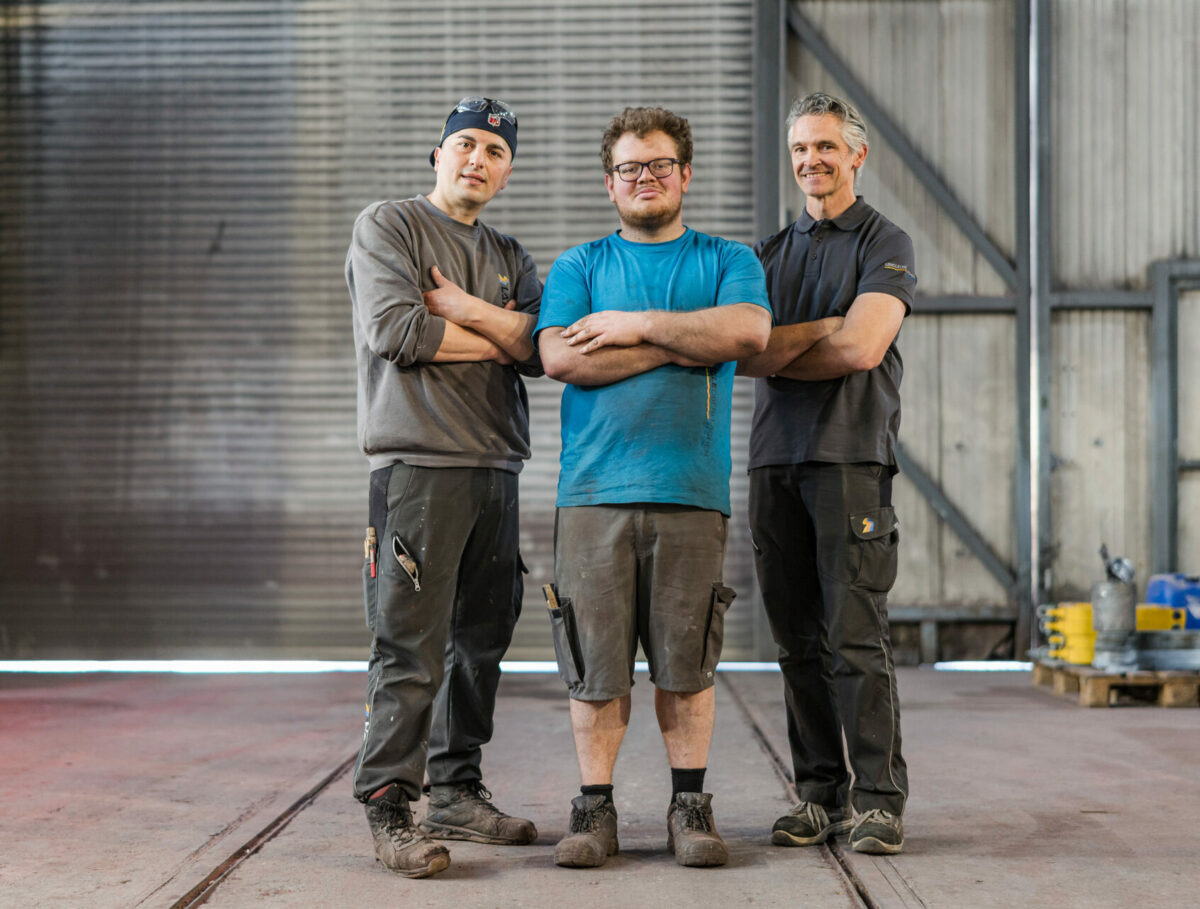 Drei Männer mit verschränkten Armen stehen in einer Industriehalle und blicken selbstbewusst in die Kamera. Sie tragen Arbeitskleidung und Sicherheitsschuhe.