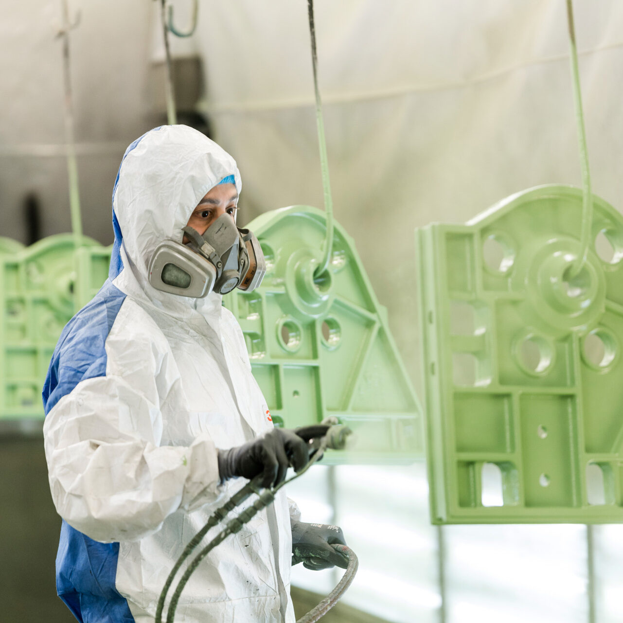 Ein Facharbeiter in Schutzkleidung und Atemmaske lackiert ein Metallbauteil in einer Lackierkabine grün.