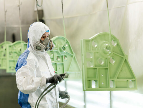 Ein Facharbeiter in Schutzkleidung und Atemmaske lackiert ein Metallbauteil in einer Lackierkabine grün.