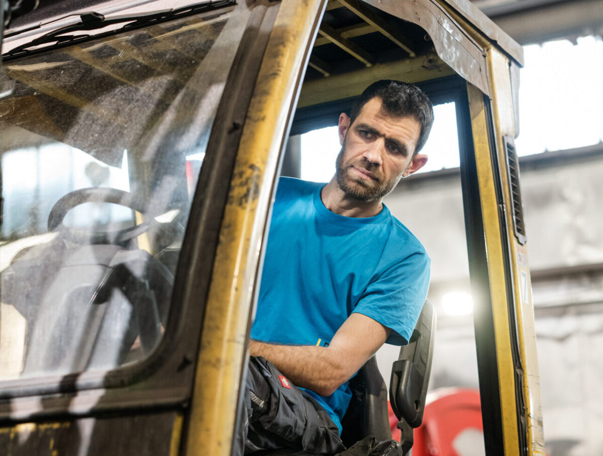 Mann in blauem T-Shirt sitzt in der Kabine eines gebrauchten gelben Gabelstaplers und schaut konzentriert zur Seite.