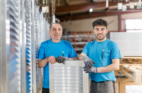 Zwei Mitarbeiter in blauen Längle-Shirts stehen lächelnd in der Produktionshalle und halten ein Aluminiumteil zum Pulverbeschichten.